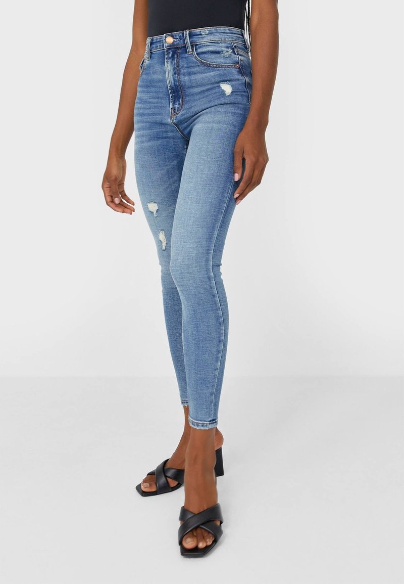 Kape skinny blue denim jeans yang menampilkan pinggang tinggi, detail rusak, dan bentuk yang pas, dipadukan dengan sandal hak tebal hitam.