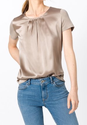 Woman wearing a beige satin short-sleeve top with pleats at the neckline and fitted blue jeans against a white background.