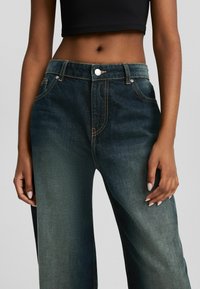 Midja på en person som bär högmidjade, mörkblekta jeanstights och en svart kort topp, med händerna vila lätt vid sidorna.