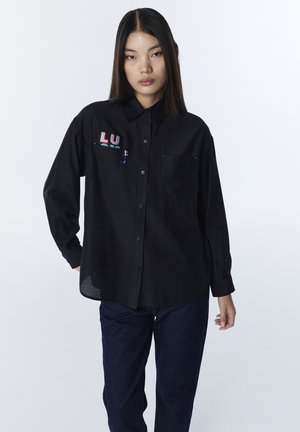 Jonge vrouw met steil donker haar, gekleed in een zwart overhemd met knopen en een kleurrijk ontwerp op het borstzakje, gecombineerd met donkerblauwe spijkerbroek.