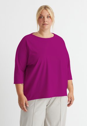 Magenta Übergrößen-Oberteil mit rundem Ausschnitt und drei Viertelärmeln, aus weichem Stoff gefertigt. Kombiniert mit hellbeigen Hosen, minimalen Texturdetails.