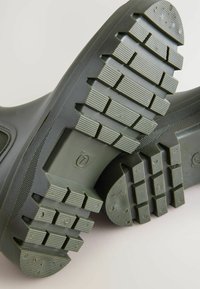 Zwarte rubberen laarzenzolen met een getextureerd profielpatroon, diepe groeven en verhoogde blokken voor grip. Stevige constructie met een lichte kromming.