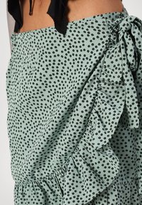 Jupe en tissu vert menthe avec un motif à pois floraux noirs, un ourlet à volants et un détail de nœud sur le côté pour la fermeture.