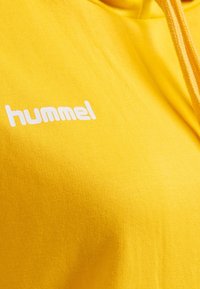 Gul hoodie av mjukt tyg, med en vit "hummel"-logotyp tryckt på framsidan, med dragsko på huvan och strukturerad yta.