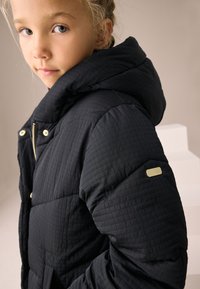 Doudoune noire à texture matelassée, grande capuche, éléments métalliques dorés et un logo accentué sur la manche. Coupe ajustée avec fermeture éclair.