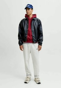 Jeune homme portant une casquette bleue, une veste en cuir noire par-dessus un sweat à capuche rouge, un pantalon de survêtement gris clair et des baskets beiges, se tenant devant un fond uni.