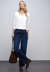 Top bianco a maniche lunghe con scollatura a V, abbinato a jeans a gamba larga blu scuro. Gli accessori includono una borsa a spalla marrone e stivaletti coordinati.
