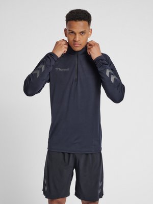 Junger Mann richtet den Kragen eines schwarzen langärmeligen Sporttops mit reflektierenden Chevron-Mustern und passenden schwarzen Shorts.