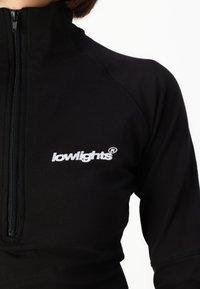 Fekete cipzáras sportfelső hosszú ujjakkal, fehér hímzett "lowlights" logóval a mellkasán és síkvarrásos kivitelezéssel.