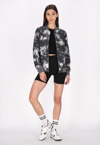 Jachetă bomber neagră și gri cu un model asemănător norilor, asortată cu un top scurt negru și pantaloni scurți negri. Adidași albi cu accente negre.