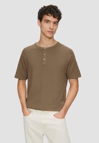 Braunes Kurzarm-Henley-Shirt mit drei Knöpfen, strukturiertem Stoff, Rundhalsausschnitt und lockerer Passform, kombiniert mit hellen Jeans.