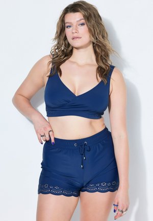 Mujer con cabello ondulado que lleva un top corto sin mangas azul marino y pantalones cortos a juego con adorno de encaje, de pie contra fondo blanco.