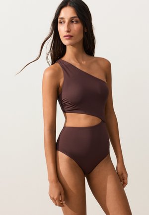 Femme portant un maillot de bain marron asymétrique avec découpe à l’épaule, debout et regardant vers sa gauche sur un fond clair uni.