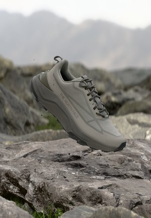 Grauer The North Face Wanderschuh mit Schnürsenkeln springt über felsiges Terrain, mit verschwommenen Bergen im Hintergrund.