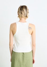 Débardeur sans manches blanc en tissu côtelé, au design ajusté, avec un col rond, affichant des tatouages visibles sur les épaules du porteur.