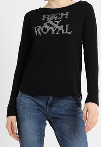 Maglietta nera a maniche lunghe realizzata in tessuto morbido, con un design "RICH & ROYAL" decorato con cristalli sul davanti. Vestibilità casual con scollo rotondo.
