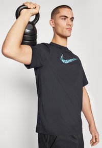 Svart atletisk skjorte med en teksturert blå og grønn Nike-logo, båret av en person som holder en svart kettlebell over skulderen.