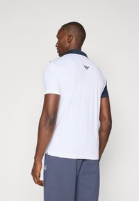 White polo shirt with navy collar and shoulder accents, featuring short sleeves and a small logo on the back. Textured fabric.
