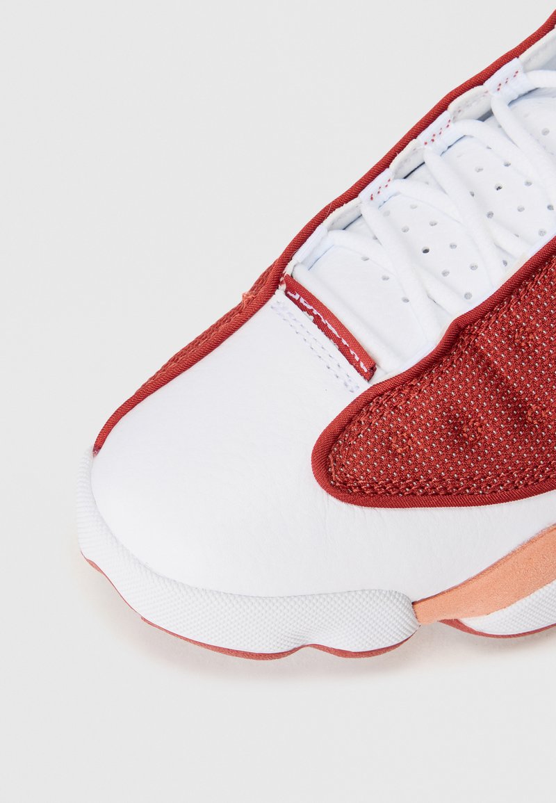 Scarpa atletica bianca con accenti in rete rossa, corpo in pelle texturizzata e lacci bianchi. Presenta una suola in gomma con un accento arancione.