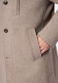 Beiger Wollmantel mit Knopfverschluss, Seitentasche und strukturiertem Stoff. Der Ärmel hat ein sauberes Ende und ein weiches Futter.