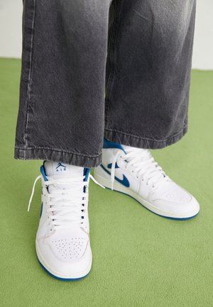 Person iført hvide og blå høje Nike Air sneakers og løse mørkegrå bukser, der står på et grønt tæppe.