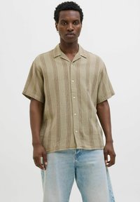 Chemise à manches courtes, col classique en vert olive clair avec un tissu texturé et des rayures brunes verticales ; portée avec un jean baggy bleu clair.
