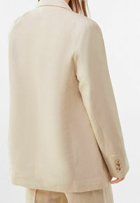 Blazer beige avec une coupe ajustée, design à simple boutonnage, col cranté et poignets à quatre boutons. Texture lisse, style minimaliste.