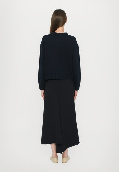 Marineblauer, kurz geschnittener Pullover mit überschnittenen Schultern, geripptem Saum und weicher Textur, kombiniert mit einem fließenden, schwarzen asymmetrischen Rock und hellen Schuhen.