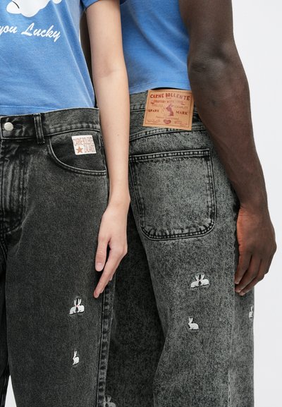 Μαύρα τζιν από denim με κεντημένα σχέδια λαγού, ορατή ετικέτα μάρκας και ξεφτισμένο τελείωμα, φορεμένα από δύο άτομα από το πλάι.