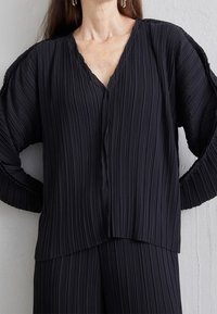 Svart plisserad topp med V-ringning och långa ärmar, med justerad textil och en asymmetrisk nederkant som ger flöde.