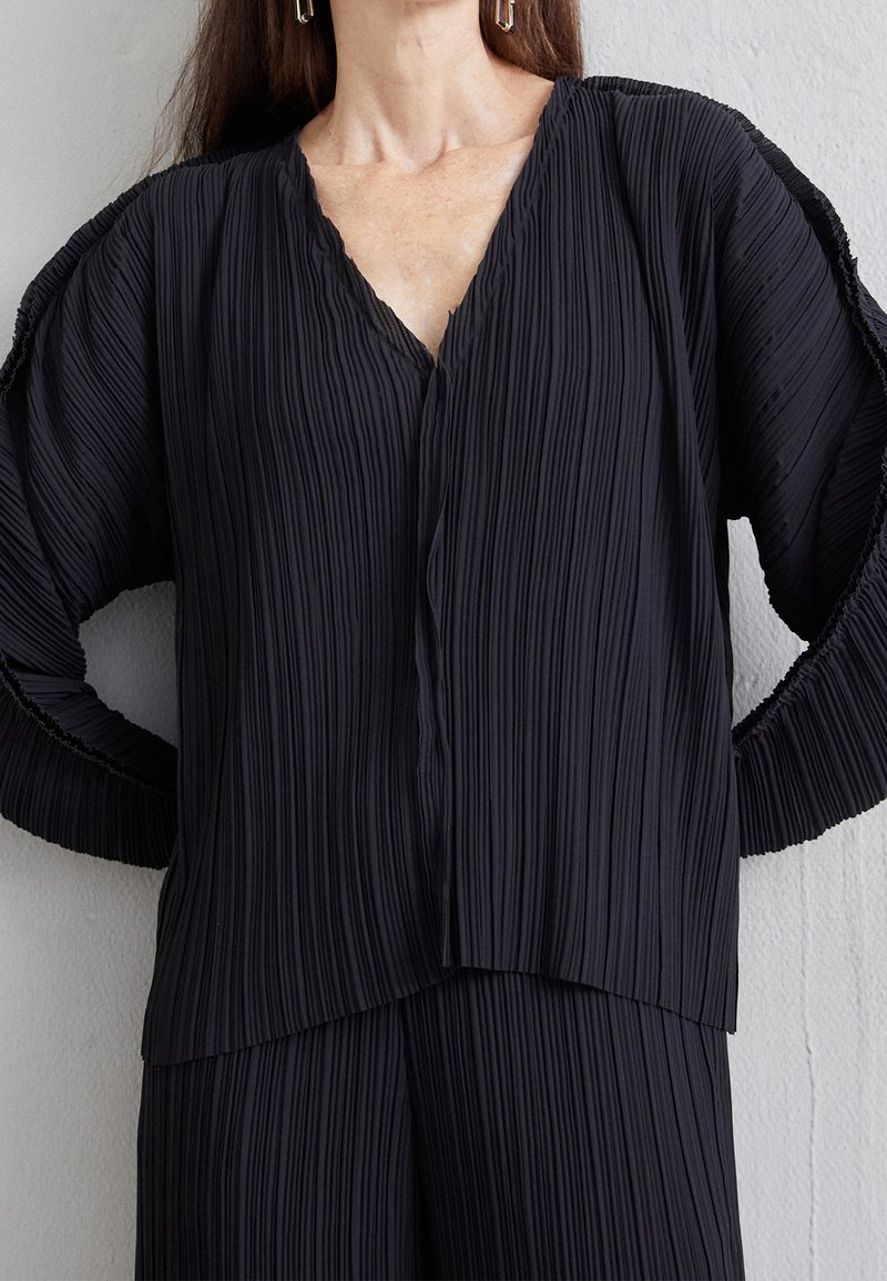 Svart plisserad topp med V-ringning och långa ärmar, med justerad textil och en asymmetrisk nederkant som ger flöde.
