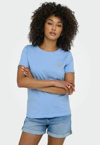 Camiseta de algodón de color azul claro con cuello redondo, mangas cortas y un detalle de corazón brillante en el lado izquierdo del pecho. Combinada con pantalones cortos de mezclilla.