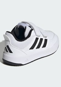 Bijela sportska cipela s teksturiranim gornjim dijelom, crnim mrežastim detaljima i tri crne pruge sa strane. Skriveni zatvarač na čičak. Gumeni đon.
