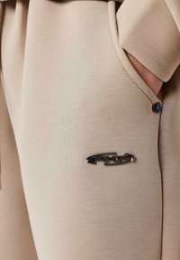 Tela de sudadera beige con textura suave, que presenta un logo metálico negro y un sutil brillo. Bolsillo lateral visible.