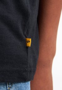 Zwarte katoenen T-shirt met een gele label met een logo, toon textuur en stikselsdetails aan de zoom. Blauwe denim zichtbaar eronder.