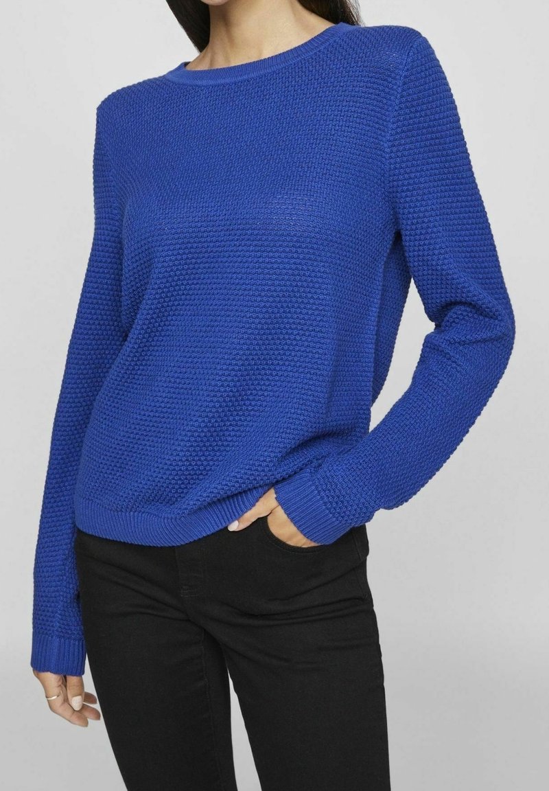 Femme portant un pull en maille texturée bleu vif à manches longues et un pantalon noir, mains dans les poches, devant un fond gris uni.