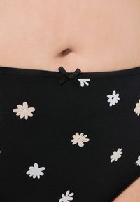 Svart underkläder med vita och beige blommönster, med en texturerad tyg och en liten svart rosett vid midjan.