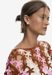 Top en crochet avec des motifs de fleurs marron, rose et crème ; fil texturé ; porté sur une base légère ; le modèle montre un profil de côté.