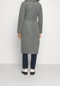Persona che indossa un cappotto grigio a spina di pesce con cintura, jeans blu scuro e scarpe bianche, in piedi di spalle contro uno sfondo neutro.