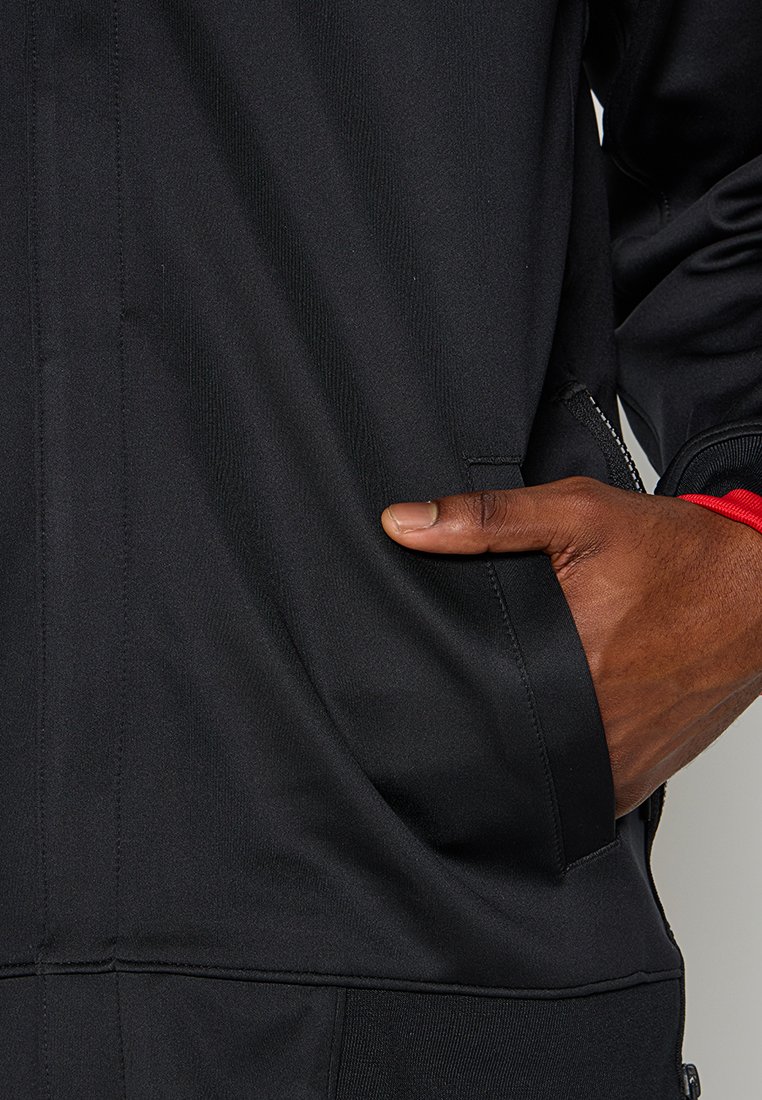Giacca sportiva nera con una texture liscia, dotata di tasche laterali con zip e dettagli rossi sui maniche.