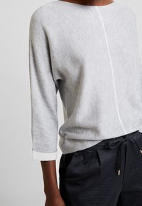 Jasnoszara bluzka z długim rękawem z miękkiego materiału, z okrągłym dekoltem i białymi detalami na szwach. W komplecie z ciemnymi spodniami w wzory.