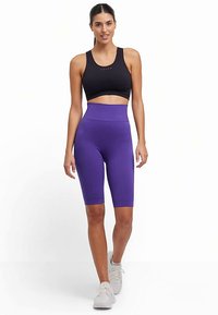 Czarny sportowy biustonosz w połączeniu z fioletowymi, wysokim stanem krótkimi spodenek rowerowych o fakturze prążkowanej. Dopełnieniem stroju są białe trampki.