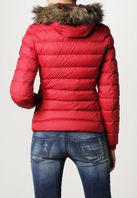 Rød pufferjakke med en aftagelig pelsforet hætte, der har et quiltet design og ribbet kant, sat sammen med blå denimjeans.