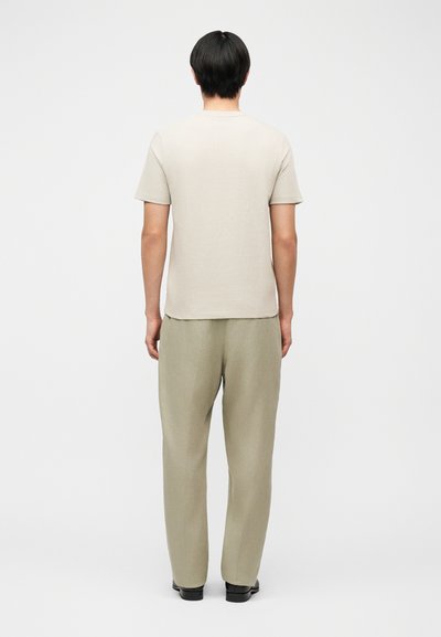 Άτομο που στέκεται με την πλάτη, φοράει ανοιχτό μπεζ κοντομάνικο πουκάμισο, φαρδιά χακί παντελόνια και μαύρα παπούτσια μπροστά σε απλό φόντο.