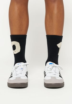 Zapatillas blancas y grises con rayas negras, combinadas con calcetines negros que tienen un diseño en punto color crema con el número 10.