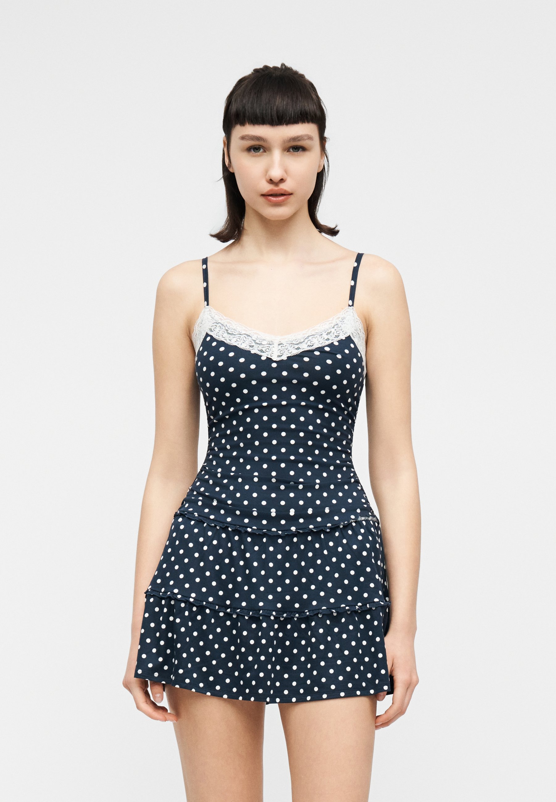 Young woman with short dark hair wearing a navy blue polka dot camisole dress with white lace trim, standing against a plain background.