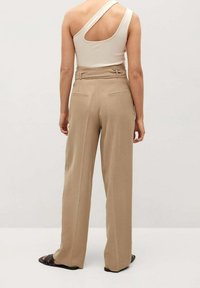 Kobieta w beżowych spodniach z wysokim stanem i szerokimi nogawkami oraz białym topie na jedno ramię bez rękawów, stojąca plecami na jednolitym tle.