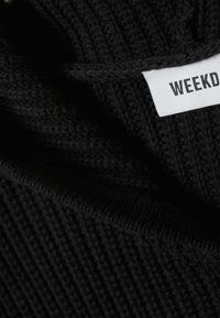 Tecido preto tricotado com textura canelada, apresentando uma etiqueta branca com a inscrição "WEEKDAY." O material parece macio e espesso.