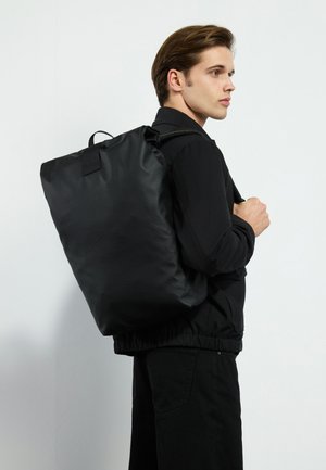 Black nylon backpack featuring a fold-over top and a carry handle, worn on the shoulder, with adjustable straps and a smooth texture.