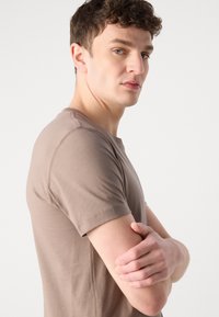 Majica kratkih rukava, smeđa pamučna majica s okruglim dekolteom i glatkom teksturom. Model prekriženih ruku, prikazuje bočni profil.